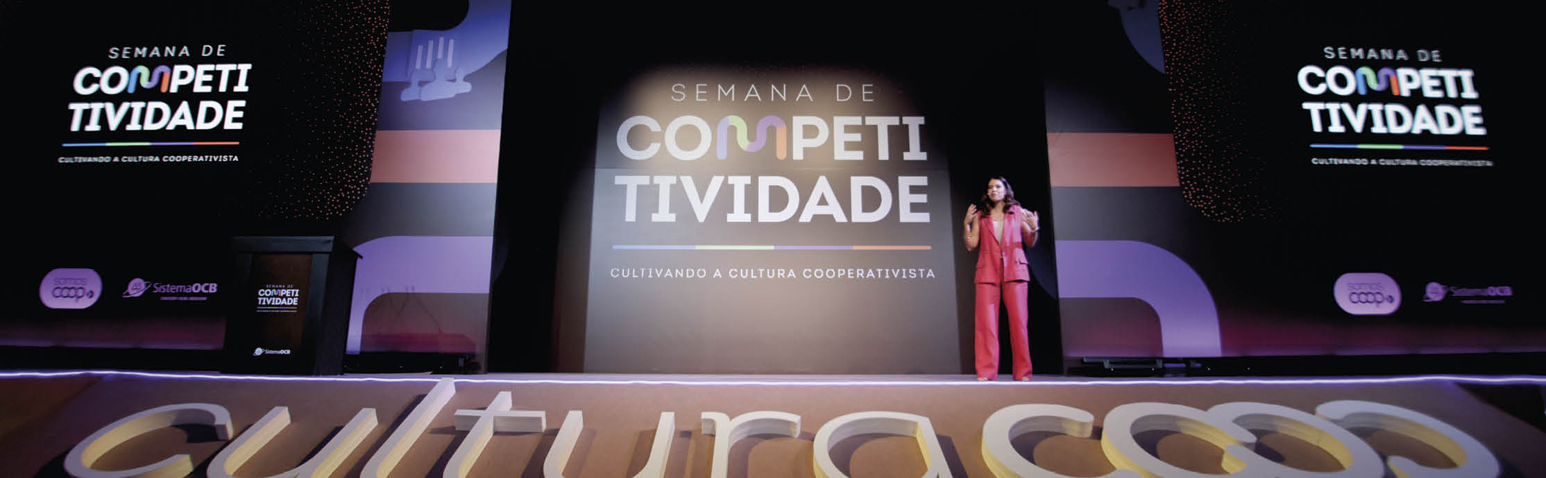 Evento chamado Semana de Competitividade com uma pessoa vestindo terno vermelho falando em um palco iluminado com telas ao fundo exibindo o nome do evento e a frase “Sustentando a cultura cooperativista". Description generated by AI