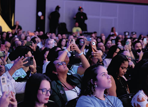 Pessoas sentadas em um audit rio lotado assistem a um evento enquanto v rias delas registram o momento com seus celulares. Description generated by AI
