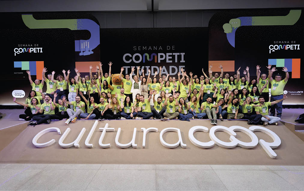 Grupo grande de pessoas vestindo camisetas verdes posando animadamente em palco com decora o e letreiro que diz cultura coop durante evento Semana de Competi.  Description generated by AI