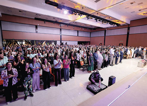 Grande grupo de pessoas em um audit rio moderno aplaudindo enquanto uma pessoa ajusta equipamentos eletr nicos no ch o pr ximo ao palco. Description generated by AI