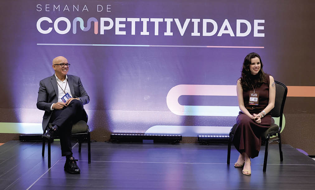 Dois adultos sentados em cadeiras em um palco durante um evento chamado Semana de Competitividade, com um fundo roxo e roupas formais. Description generated by AI