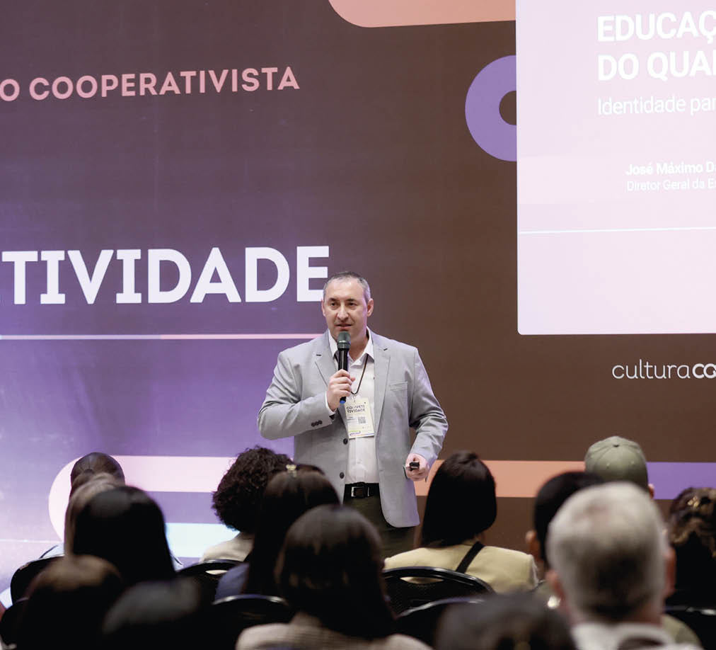 Pessoa adulta vestindo blazer cinza fala ao microfone para uma plateia sentada em evento com apresenta o projetada ao fundo.  Description generated by AI