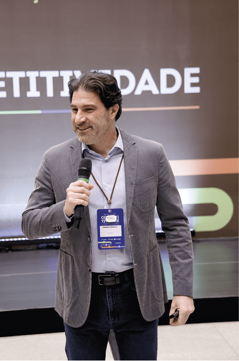 Pessoa adulta com cabelo ondulado e escuro segura microfone e crach em evento com fundo escuro e texto parcialmente vis vel.  Description generated by AI