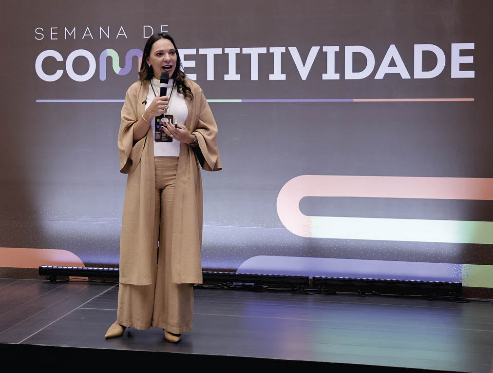 Pessoa adulta com cabelo longo e solto vestindo conjunto bege fala ao microfone em palco com fundo escrito Semana de Competitividade. Description generated by AI