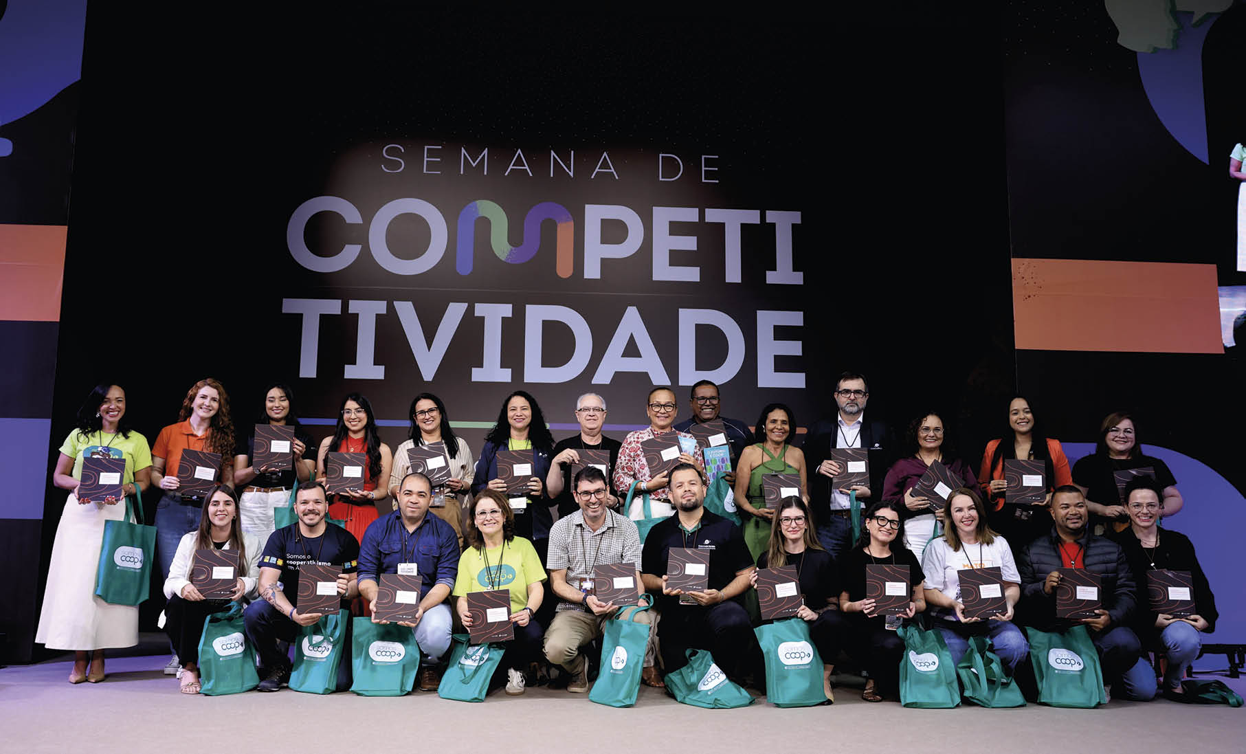 Grupo de adultos sorridentes segurando placas e bolsas verdes em evento com painel ao fundo escrito Semana de Competitividade. Description generated by AI