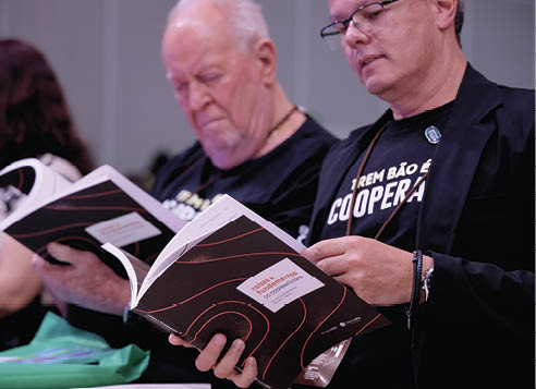 Duas pessoas sentadas lendo livros com capa preta e detalhes em vermelho em um ambiente interno. Description generated by AI
