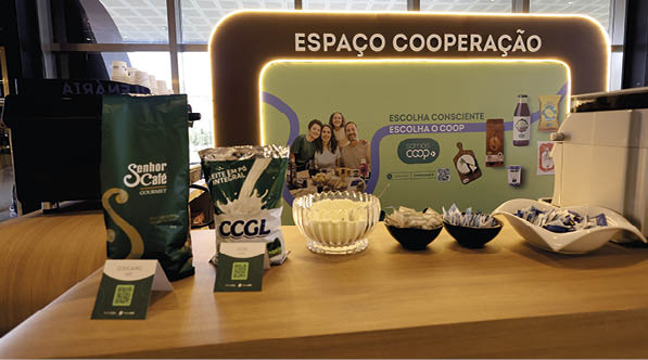 Espa o de coopera o com produtos de caf  expostos sobre uma mesa de madeira e um painel iluminado ao fundo com pessoas sorrindo e mensagens sobre escolha consciente.  Description generated by AI