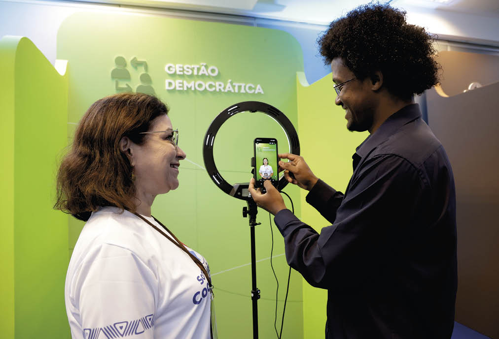 Duas pessoas interagem em um estande com fundo verde escrito “Gest o Democr tica", uma segura um celular diante de um anel de luz enquanto a outra sorri olhando para o aparelho. Description generated by AI