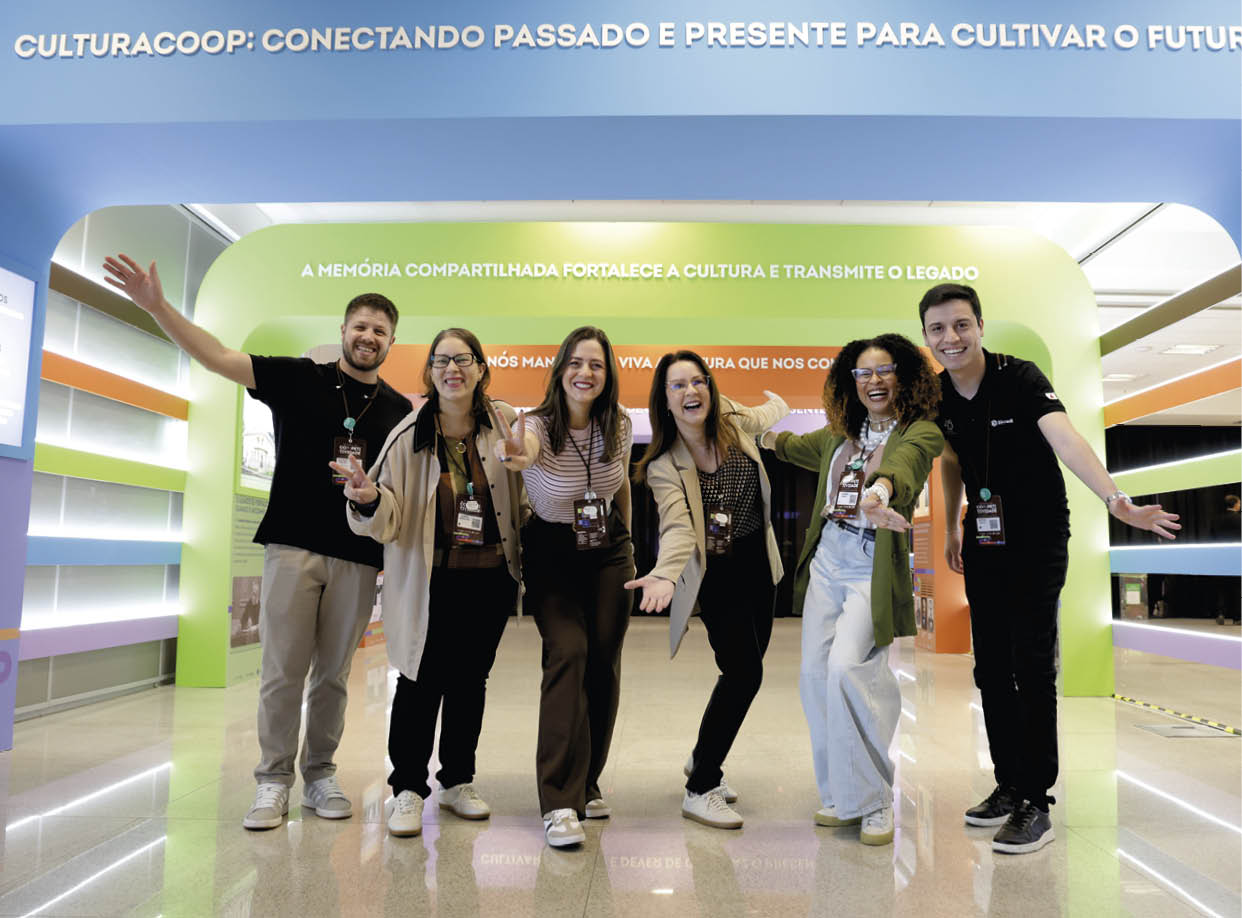 Seis pessoas adultas sorridentes est o posando com os bra os abertos sob uma estrutura azul com texto em portugu s em um ambiente interno iluminado. Description generated by AI