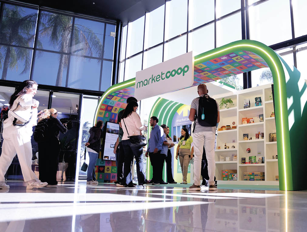 Pessoas conversam e observam produtos em um estande colorido com o letreiro “marketcoop" em um ambiente interno iluminado por grandes janelas. Description generated by AI