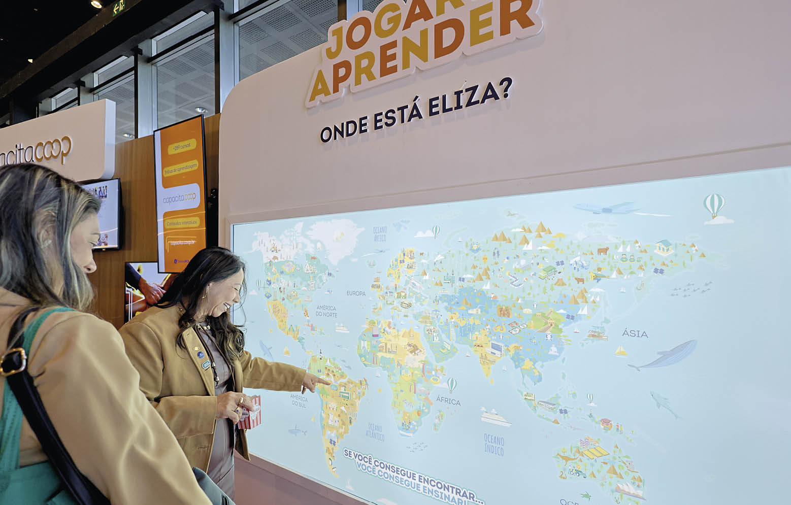 Duas pessoas observam e apontam para um mapa-m ndi interativo em uma exposi o com o t tulo “Jogar Aprender" e a pergunta "Onde est  Eliza?" escrita acima do mapa.  Description generated by AI