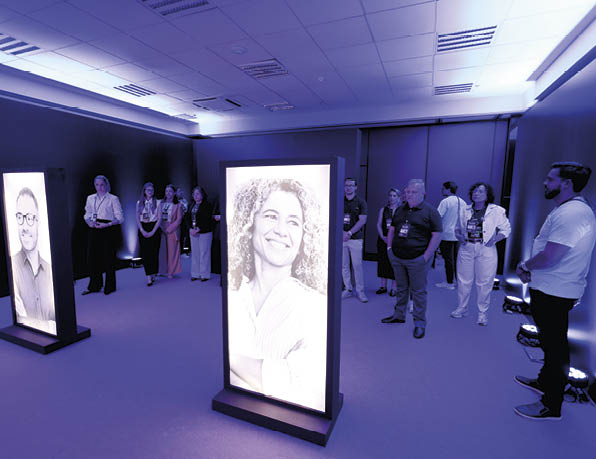Pessoas em um ambiente interno observam duas telas iluminadas com retratos em preto e branco de pessoas sorrindo. Description generated by AI