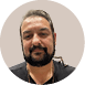 Pessoa adulta com barba e cabelo curto penteado para tr s usando camiseta preta e colar dourado, com fundo neutro circular. Description generated by AI