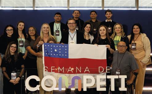 Grupo de pessoas sorrindo em ambiente interno segurando uma bandeira com as palavras “Semana de Competi o".  Description generated by AI