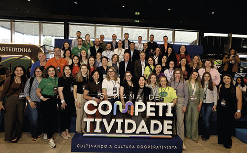 Grupo grande de adultos reunidos em ambiente interno posando para foto durante evento chamado Semana de Competitividade, com destaque para um letreiro colorido e uma faixa com a frase Cultivando a cultura cooperativista. Description generated by AI