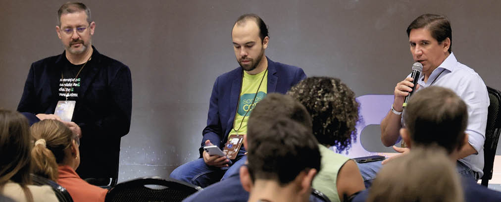 Tr s adultos sentados em cadeiras no palco participam de um painel enquanto v rias pessoas assistem em uma sala escura. Description generated by AI